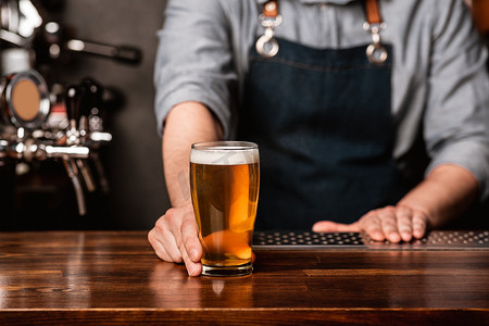 在酒吧发放啤酒。穿围裙的酒吧招待在木制柜台上给顾客一杯淡啤酒