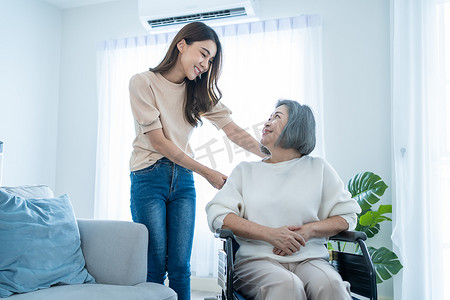 Asian young daughter support older senior woman on wheelchair at home. Beautiful girl granddaughter help and take care of elderly mature mother patient doing physical therapy in living room in house.