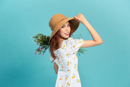 架势摄影照片_身穿夏装，头戴草帽的年轻美丽的亚洲女人，孤零零地站在蓝色的背景上，摆出一副花束的架势