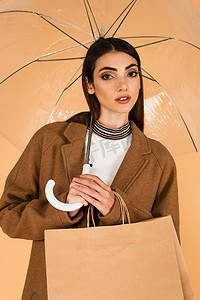 穿着时髦的女人站在伞下看着相机，手里拿着用米黄色隔开的购物袋