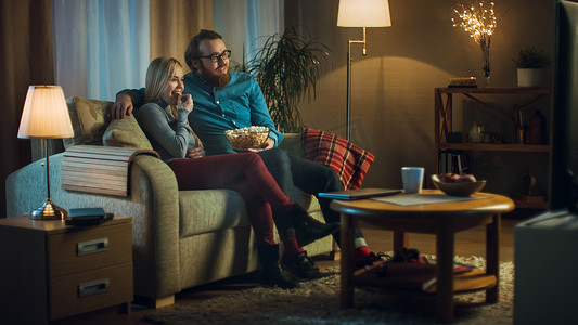 电视墙造型摄影照片_晚上男人和女人坐在沙发上看电视，吃爆米花。客厅是舒适.