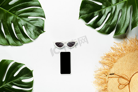 手机绿色背景摄影照片_绿色棕榈叶、草帽、太阳镜和带有智能手机白色背景的记事本的顶部视图