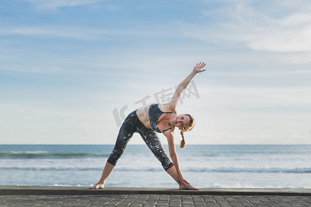 妇女练习瑜伽在扩展三角姿势与海洋和蓝天背景下, 巴厘岛, 印尼