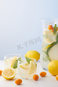 壶和两杯柠檬水与薄荷叶, 冰块和柠檬片周围的金橘和柠檬在蓝色背景下 