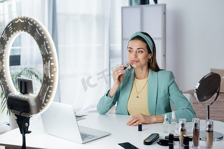 pretty woman applying lipstick near laptop, smartphone with blank screen and circle lamp