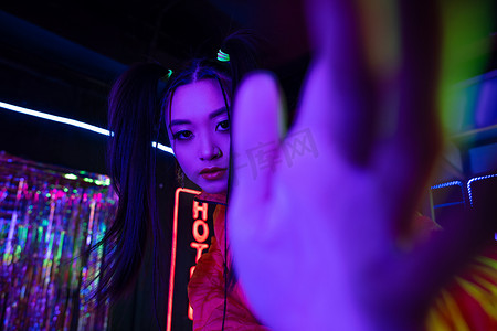 young asian woman with blurred hand near neon sign with hotel lettering outside 