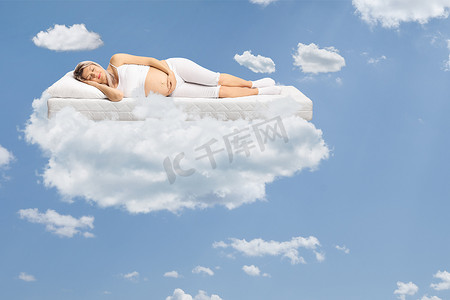 pajamas摄影照片_Pregnant woman in pajamas sleeping on a mattress and floating on clouds in the sky