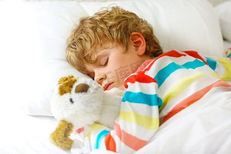 可爱的金发碧眼的小男孩穿着五颜六色的睡衣，睡在白色的床上，带着玩具做梦。健康的孩子，有柔软的玩具，安安静静地睡在家里