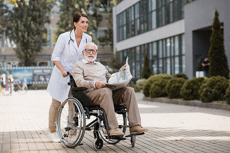 老年护士与坐在轮椅上的老年残疾人一起在户外与报纸同行