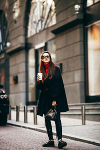 年轻的时尚女孩，路过 Windows.Wearing 时尚眼镜和一件黑色外套。可以使咖啡.