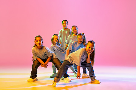 Joy, fun and happiness. Portrait of cheerful, active little girls, happy kids dancing isolated on orange background in neon light. Concept of music, fashion, art, childhood, hobby.