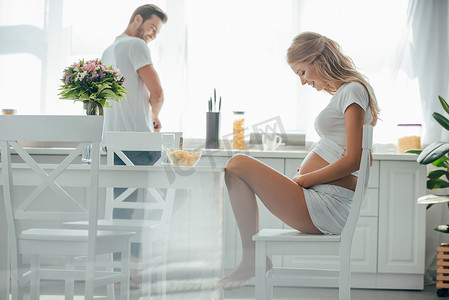 坐在餐桌上的孕妇与水果沙拉在碗和丈夫在厨房柜台的侧面视图