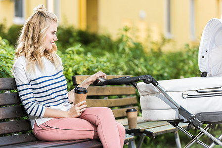 微笑的母亲坐在长凳上的侧面观看咖啡去和举行婴儿推车在公园里