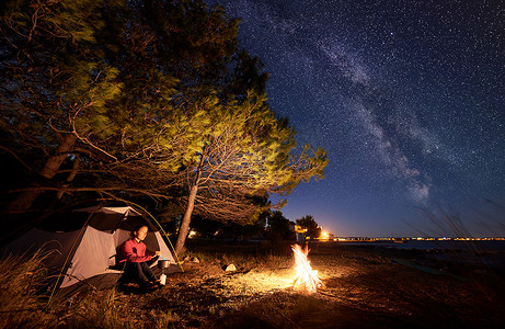 银河星光摄影照片_在海边过夜露营。徒步旅行的妇女准备食物在一个气体燃烧器, 休息在旅游帐篷入口在森林附近对明亮的蓝色星光天空与银河