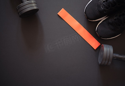 红色弹性橡皮筋用于健身，黑色运动鞋和黑色哑铃用于黑色背景的运动。一套运动器材。适合在家里训练。案文的篇幅.