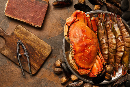 在碗中未煮熟的螃蟹、贝类、公鸡和贝类的顶视图，靠近碎裂的旧书和在纹理表面的木切碎板上的海鲜饼干 