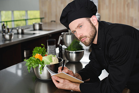 英俊的厨师写下新食谱的笔记本在餐厅厨房和看着远离