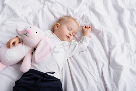 被褥摄影照片_俯瞰幼儿与玩具兔子睡在白色被褥上的景象