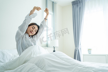 身穿睡衣的亚洲美女早上醒来，心情很愉快。迷人的年轻女子笑着、快乐着、放松着，然后从床上躺在家里的床上舒展身体.