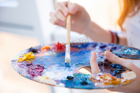 paints摄影照片_Paintbrush in woman hands mixing paints on palette
