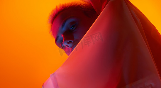 From below young female model looking at camera from behind futuristic apparel while standing under red neon light against orange background