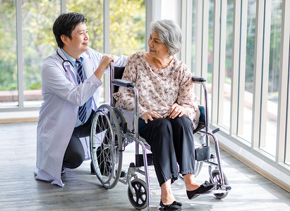 Asian male professional doctor in lab coat with stethoscope helping pushing pointing outside view to happy old senior gray hair retired pensioner handicapped patient sitting smiling on wheelchair.