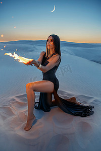 一个神秘的女人坐在沙滩上，手里拿着一把火。幻想，艺术照片。女孩把脸藏在金色的面纱后面.女魔头的形象。沙漠之夜的背景有缝隙的创意服装