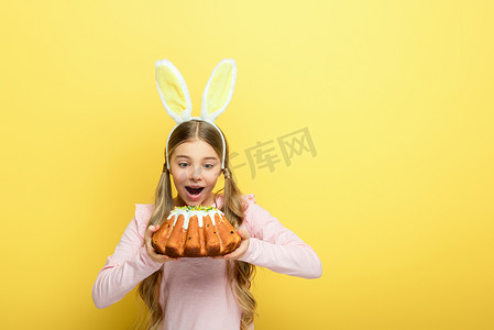 shocked kid with bunny ears holding easter cake isolated on yellow 