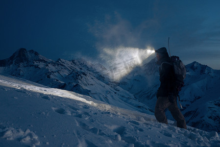 专业的特派员承诺在夜间爬上雪山, 用车头灯照亮道路。穿着滑雪服, 背包和后面的滑雪板。野外和滑雪旅游