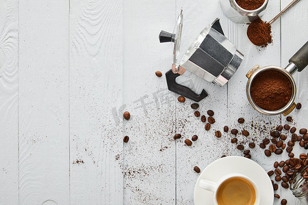 咖啡机和咖啡豆摄影照片_在白色木质表面与咖啡豆一起享用的门廊、勺子和咖啡杯附近的间歇泉咖啡机的顶视图