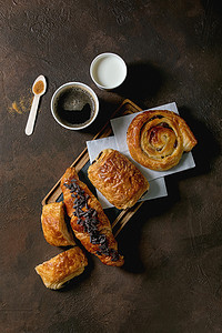 各种传统的法国松饼面包，带有拉辛和巧克力，羊角面包与咖啡和牛奶的纸杯，回收木勺甘蔗糖在黑暗的纹理背景。平坦的地方，空间