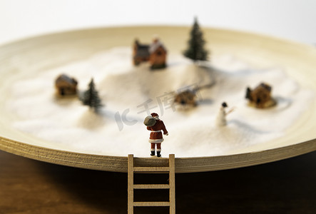 木板雪摄影照片_ 圣诞老人站立在木板材边缘与木梯子和小村庄看法在白色雪