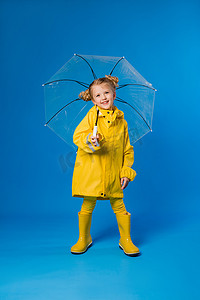 放置图片摄影照片_穿着雨衣和橡胶靴的快乐女孩站在蓝色的背景上，带着一把透明的雨伞。复制空间图片并放置到您的广告文本中
