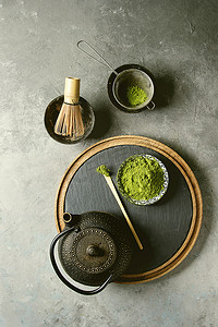 黑茶壶摄影照片_做旳饮料的配料。绿茶旳粉在陶瓷碗, 传统竹勺和扫石板板, 黑铁茶壶的灰色纹理背景。平躺, 空间