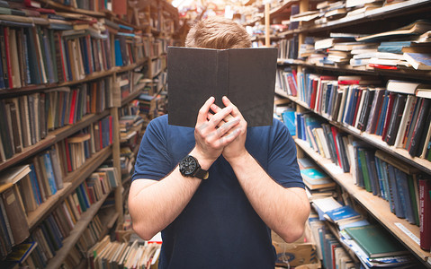 站在图书馆里的一个人的肖像用一本公开的黑色书遮住了他的脸。一个在舒适的公共图书馆手中藏在书后的男人.