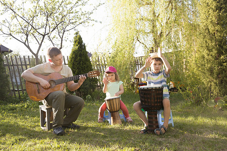 快乐的家庭爸爸和两个孩子在户外一起玩乐器。爸爸弹吉他，孩子们玩民族鼓。隔离。音乐概念