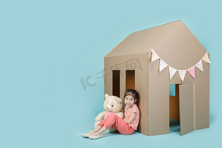 和你一起玩摄影照片_亚洲小女孩和她的玩具熊一起玩纸板屋，用蓝色的长横幅隔开，上面有抄袭的空间，作为你的课本，带着家庭观念在家里创作