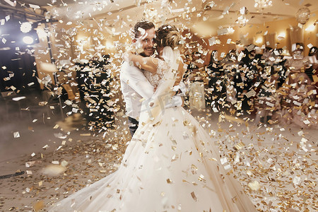 华丽的新娘和时尚的新郎在婚礼招待会的金色五彩纸屑下跳舞。快乐的新婚夫妇在餐厅表演第一支舞。浪漫时刻