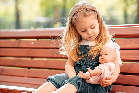  金发小女孩在公园里散步，在长椅上玩洋娃娃，做母亲的女儿游戏，夏天阳光灿烂的日子，快乐孩子日的概念