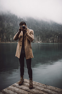 湖畔小提琴摄影照片_全身上下都是身穿棕色外套的年轻男性摄影师，他站在奥地利普莱西湖畔的湖畔小屋甲板上，用一个小的无镜像相机拍照