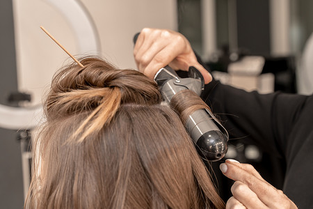 理发师准备一个女人，用卷发用卷发熨斗做发型。长长的浅棕色自然头发。自我照顾的概念.