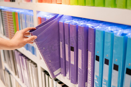 民生用品摄影照片_男性手选择新的紫色环活页夹文件夹从五颜六色的货架显示在文具店。购买办公用品概念