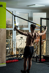 trx摄影照片_体育和健身房的概念。男子与躯干, 运动员, 运动员, 肌肉男子气概的运动与 trx 循环, 窗口的背景。男子裸躯干, 肌肉背部在健身房享受训练, trx