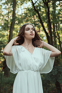 年轻温柔的妇女摆在白色礼服和花卉花圈在森林里
