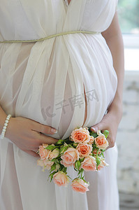 在怀孕的妇女，戴着长长的白色连衣裙，手束玫瑰鲜花拿肚子上特写