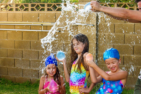 水袋气球摄影照片_父亲的胳膊在他的三个女儿的头上打碎了一个蓝色的水气球。女孩们穿着泳衣, 其中两人有游泳帽上.