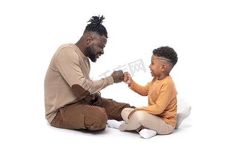 黑皮肤的非洲男人和他的小儿子在一起。他们坐在地板上，在工作室的白色背景下玩着有趣的游戏
