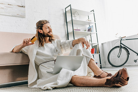 微笑的耶稣坐在地板上用信用卡和做网上购物在膝上型电脑附近沙发在家