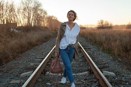 时尚生活方式的肖像年轻的时尚妇女穿着棕色外套, 白衬衫和牛仔裤摆姿势, 笑着走在火车轨道上回家的村庄与森林在秋天。愉快的妇女的画像