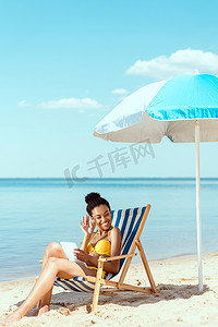 非洲裔美国人妇女用数字片放松在甲板椅子和手挥动在海滩伞之下在海前面 